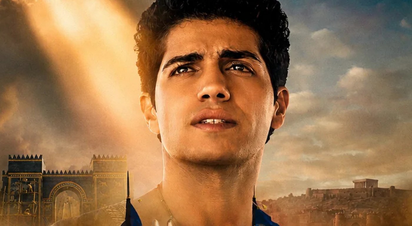 Mena Massoud as Daniel looks upward toward a shaft of light, with Babylon’s blue Ishtar Gate and a hillside city behind him