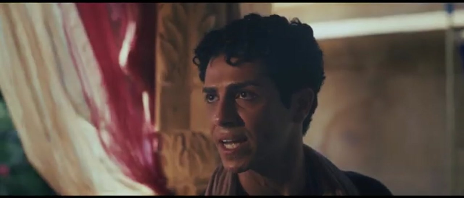 Mena Massoud as Daniel in a close-up, looking aside with concern beside pale curtains and carved stone in a palace interior