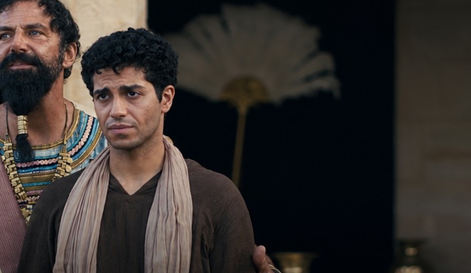 Elijah Alexander as Nebuchadnezzar rests a hand on Mena Massoud as Daniel’s shoulder inside a palace chamber in Daniel and the Fiery Furnace