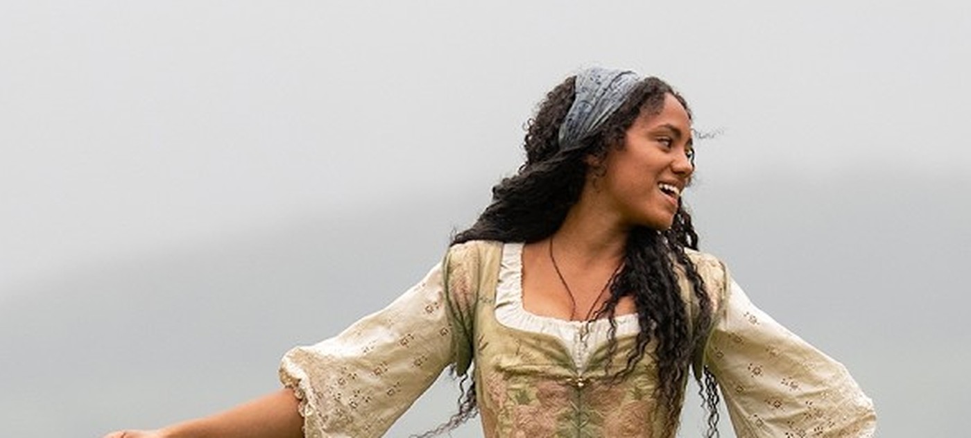 India Brown as Emmaline smiles with arms open in a cream blouse and floral bodice on a misty hillside in Drummer Boy