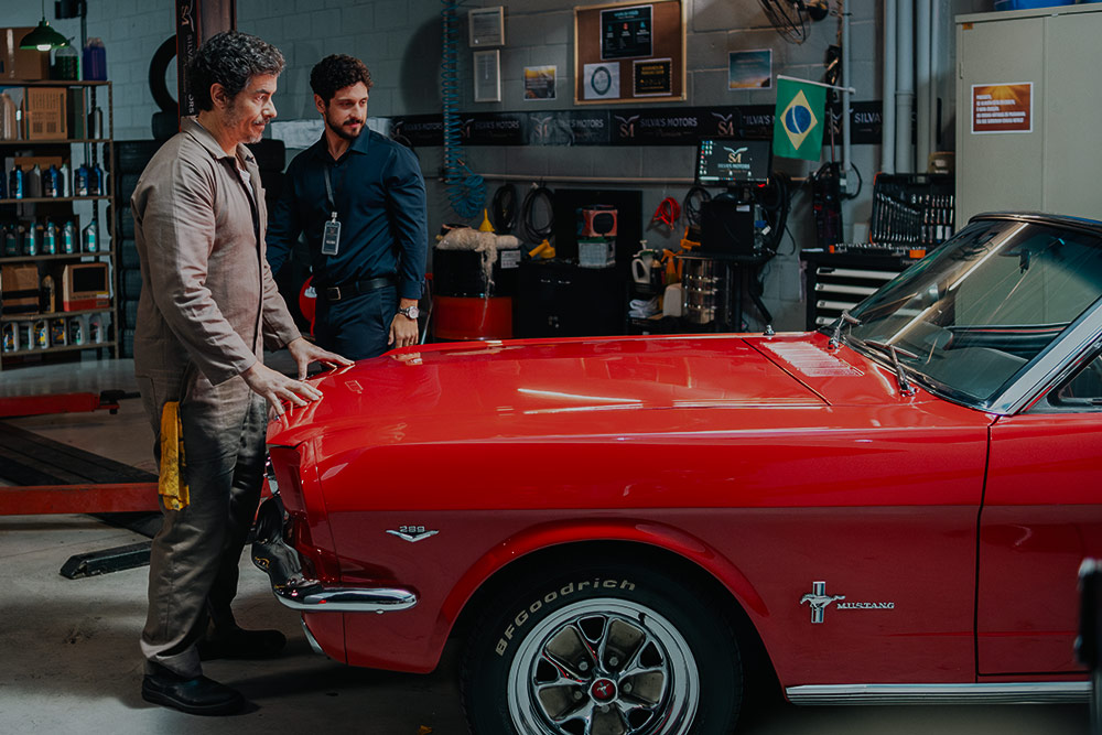 Two men stand beside a red Ford Mustang in a garage marked Silva Motors, a Brazilian flag on the wall, in Flywheel: Ignition of the Soul