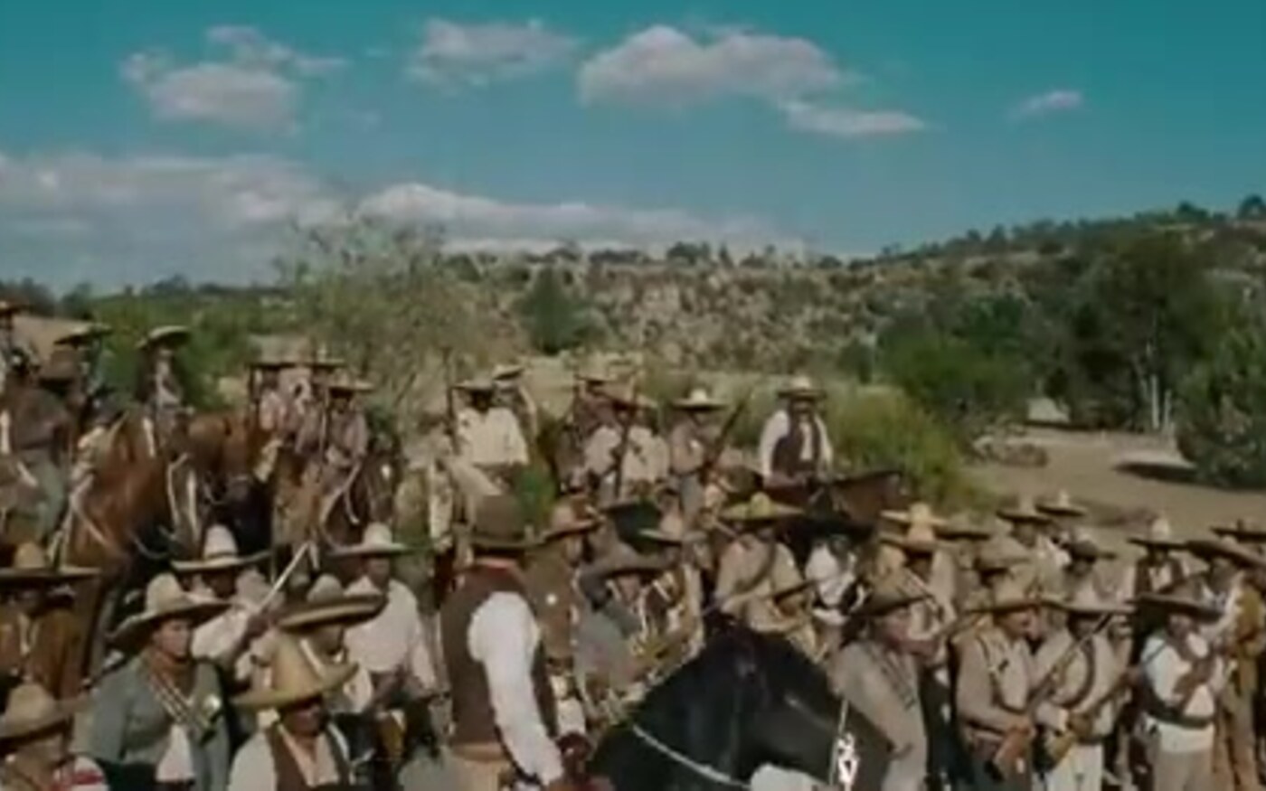 Mounted fighters ride past colonial buildings and hills in a frame from For Greater Glory